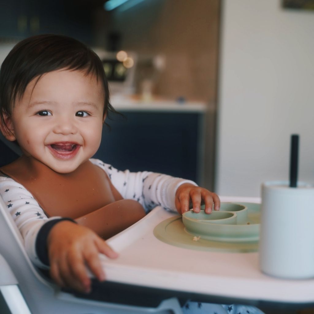 Baby boy sitting in a highchair enjoying lunch with his feeding gear from the Family Flavors Mealtime Kit. The EZPZ mini mat makes self-feeding easy and fun!